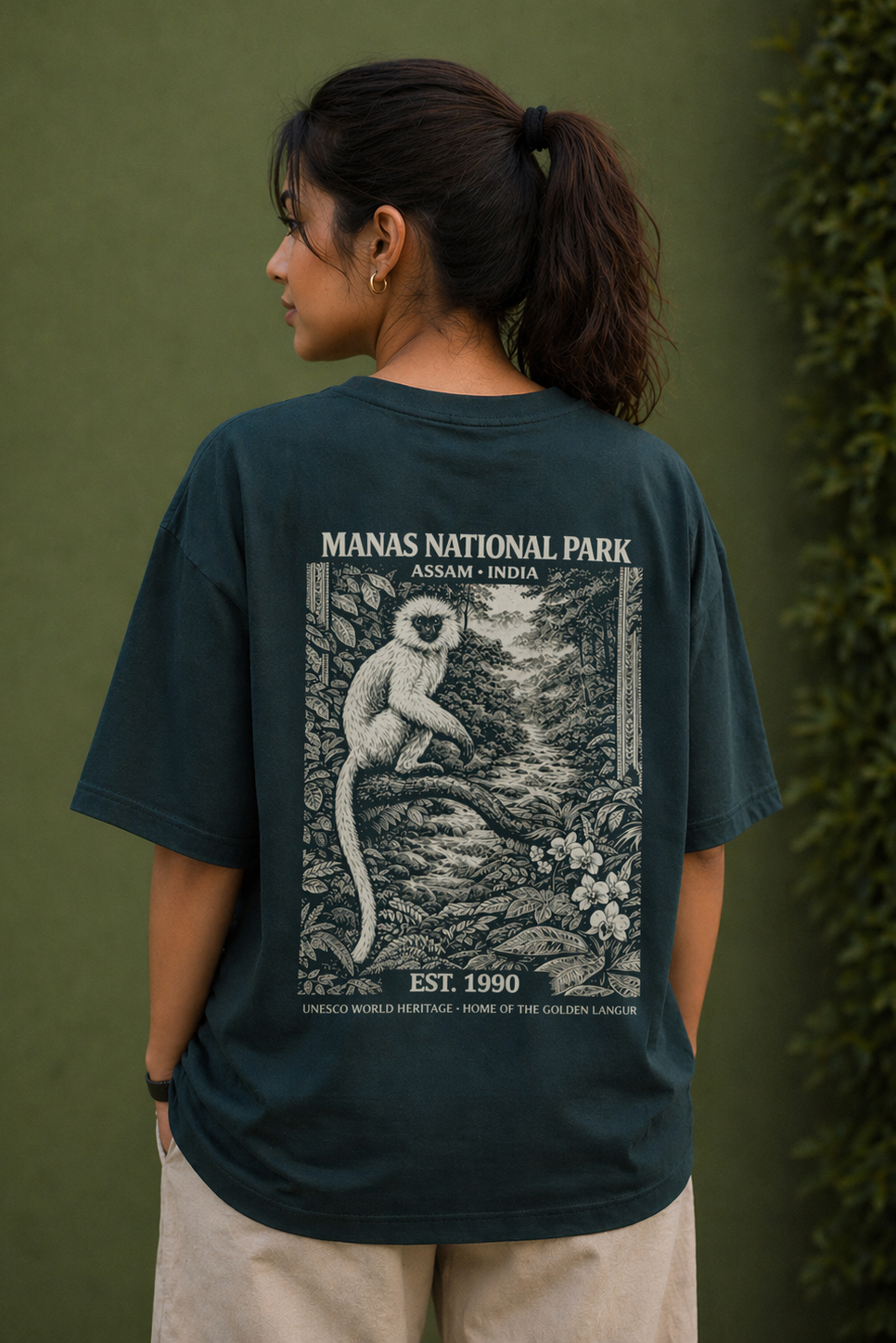 Person wearing a dark green t-shirt with Manas National Park graphic on the back, standing against a green background.
