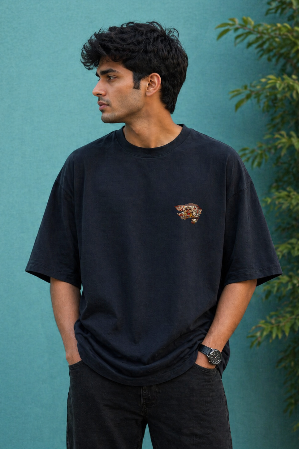 Man wearing a black t-shirt with a small logo against a teal wall with greenery.