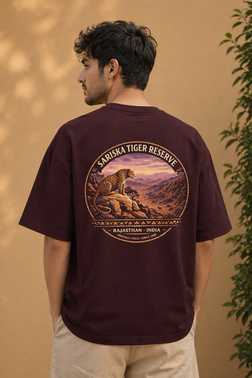 Person wearing a maroon t-shirt with a graphic of a leopard and text on the back, standing against a beige wall.