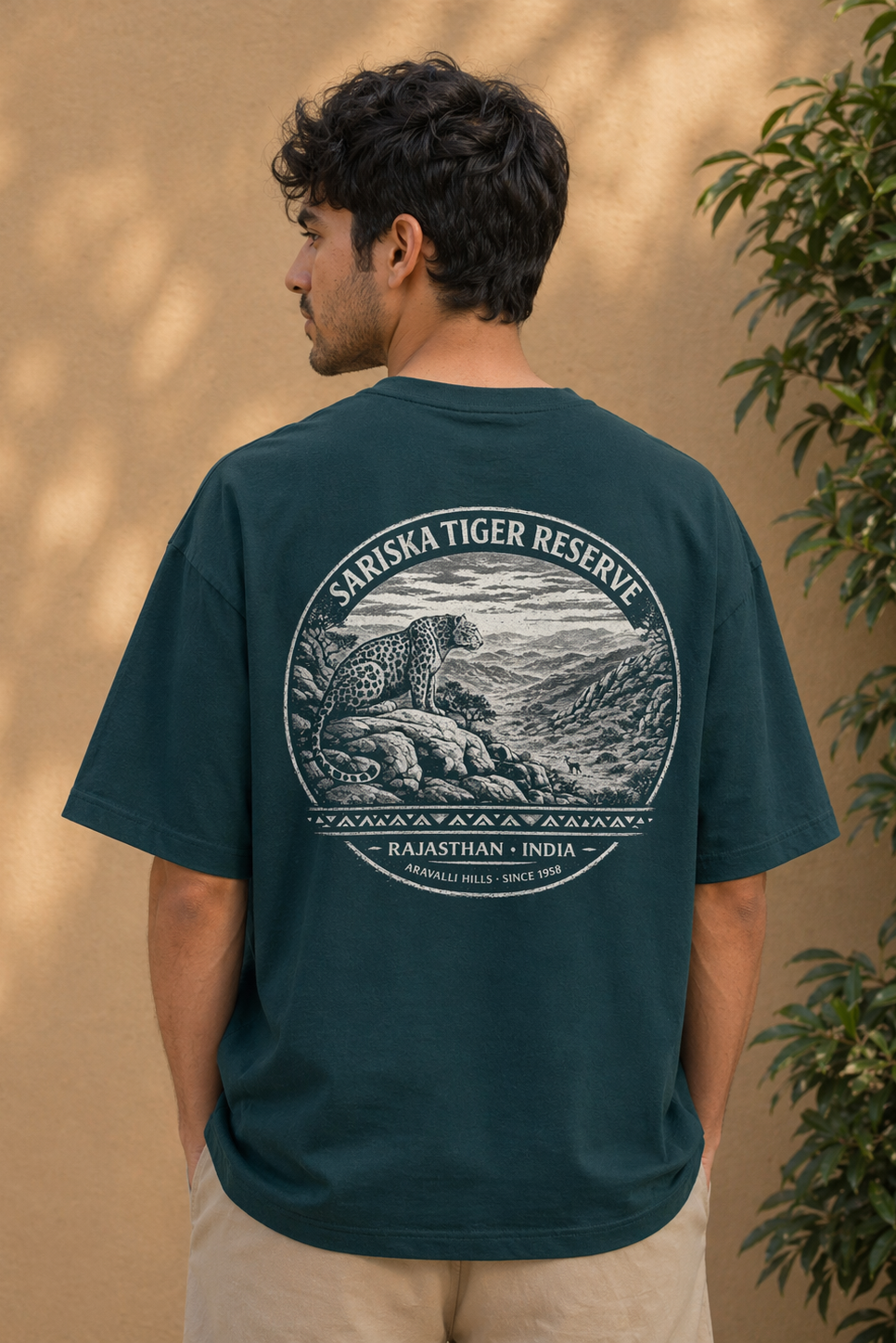 Person wearing a dark green t-shirt with a graphic of a tiger and text on the back, standing against a beige wall with plants.