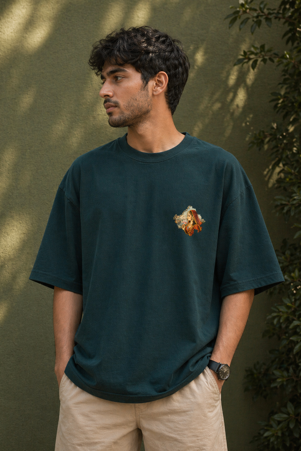 Man wearing a dark green t-shirt with a small graphic design, standing against a green wall with foliage.
